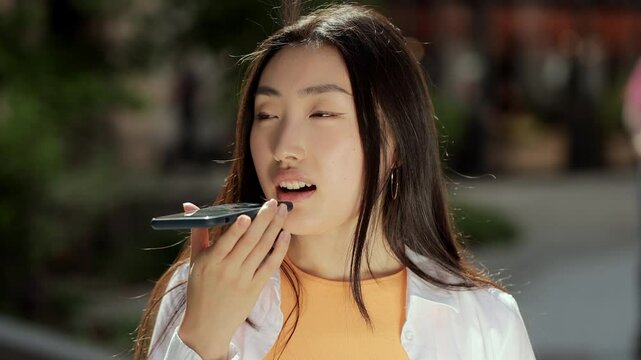 Happy young asian woman sending audio voice message on cellphone at outdoor talking to mobile assistant. Mobile software concept voice search.