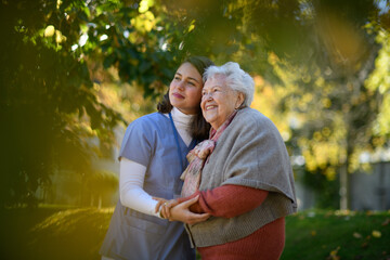 Portrait of nurse and elderly woman on walk in the park during warm autumn day. Young caregiver...