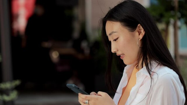 Young asian girl walking down the street while using her smartphone looking around with a smile. Modern devices, gadgets, addiction, being online. Social networks.