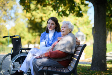 Portrait of nurse and elderly woman sitting on bench in park during warm autumn day. Young caregiver spending time with senior patient.