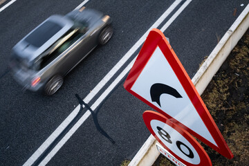 A gray car moves along the road next to a huge caution and speed limit sign. The ground is wet from the rain and there is a lot of contrast between black and red