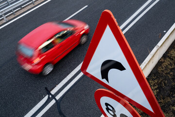 A small red car moves along the road next to a huge caution and speed limit sign. The ground is wet from the rain and there is a lot of contrast between black and red