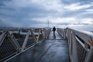 An anonymous person walks on an elevated bridge to cross the highway. It's a rainy day and the sky looks dramatic with looming clouds. There are reflections on the wet floor © LaMorenita