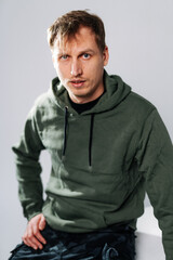 Young man in a hoodie and shorts indoors. A young man sits casually, wearing a green hoodie and patterned shorts, showcasing a relaxed and confident demeanor.