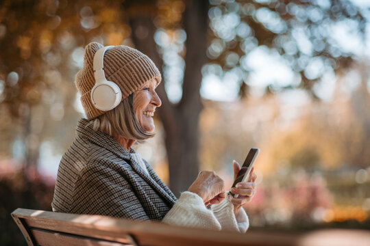 Portrait of a beautiful senior woman with headphones in autumn park, listening music and holding her phone.