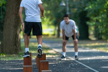 Boy balances across wooden stumps outdoors with guidance from a trainer. The activity promotes coordination, agility, and balance as part of outdoor sports exercises for children.