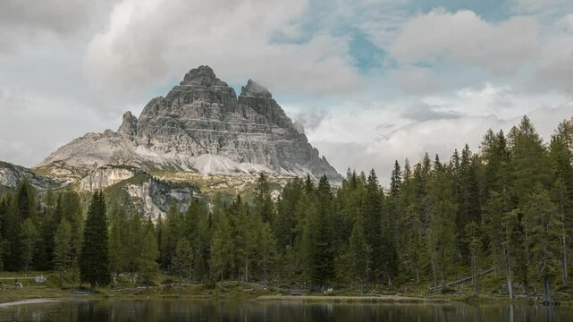 Timelapse of an awe-inspiring mountain range towering over a dense forest and lake. The rugged, rocky mountain, etched with natural ridges, stands majestically beneath a partly cloudy sky.