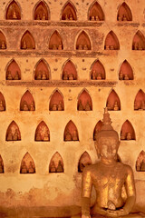A big golden, gilded buddha statue, sculpture in front of a wall filled with many miniature buddha figurines in the cloister wall of Wat Si Saket, a buddhist temple in Vientiane, Laos