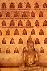 A golden, gilded buddha statue, sculpture in front of a wall filled with many miniature buddha figurines in the cloister wall of Wat Si Saket, a buddhist temple in Vientiane, Laos