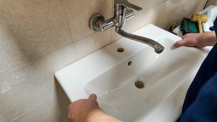A Caucasian plumber installs a white ceramic sink against a beige tile wall, addressing home repair and maintenance issues
