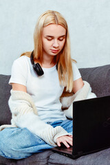 Naklejka premium A young woman works in the living room, sitting on the sofa with headphones and a laptop. A cozy moment that highlights the comfort of remote work and focus on tasks.