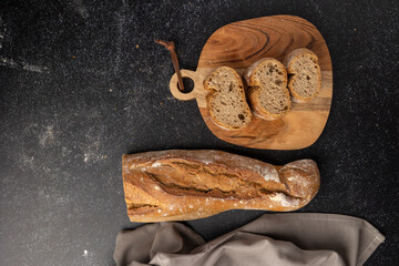 A rustic loaf of bread is placed on a wooden cutting board accompanied by a small bowl of flour, set against a dark textured background with a fabric piece to the side.