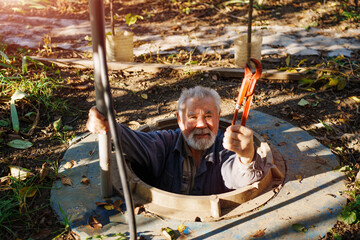 An elderly man with an adjustable wrench in the manhole of a water well. repair of water pipes.