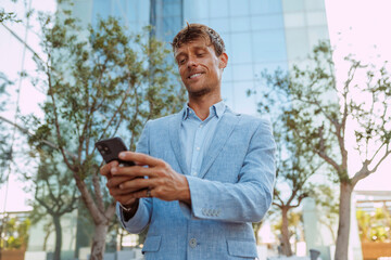 Fototapeta premium Young smiling confident attractive businessman uses smartphone outdoors standing near office building. Executive, entrepreneur, manager, men at work, technology concept.