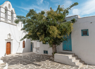 A picturesque alley in Paros features a traditional whitewashed house with bright blue doors, surrounded by quaint architecture and a lush tree casting a cool shade in the sun.