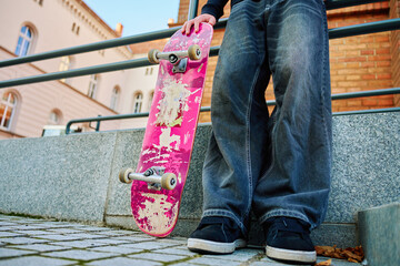 Skateboarder in baggy jeans holding scratched pink skateboard at city street. Teenager is...