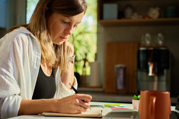 Freelance woman sitting at kitchen table using laptop and writing in notebook. Remote work at home office