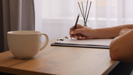 A mug on a table with a woman in the background writing in a notebook. Her writing captures the essence of thoughtful note taking. Suitable for creative themes and quiet moments of letter writing.