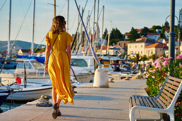 Woman is walking alone on sunny promenade with palm trees and flowers. Female tourist strolls along marina with yachts at sunset in coastal town © Lazy_Bear