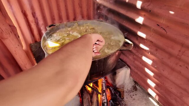 Close-up of a traditional pot on a countryside fire in the Dominican Republic, cooking Sancocho. Steam rises as a metal spoon stirs the meats and vegetables in this authentic dish.