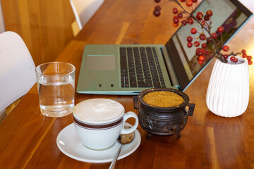 Break at work. Home office scene. A notebook, a glass of water, a cup of cappuccino, a sugar bowl and a small vase with a branch of the ornamental apple (Malus floribunda) on a wooden desk