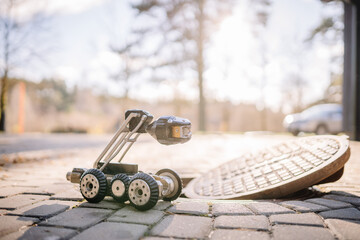 A wheeled inspection robot with a camera sits near an open manhole on a cobblestone surface, ready for underground exploration and maintenance tasks..