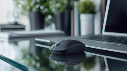 Close-up of a wireless mouse beside a laptop on a glass desk, with office plants and files in the background