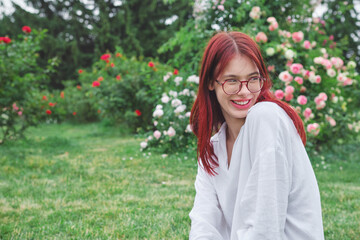 Teenage girl with red hair and glasses smiling shyly while sitting in a blooming garden on a bright day