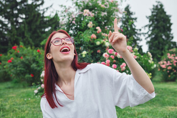 Teenage girl with red hair and glasses in a blooming garden, looking up and pointing with a surprised expression, noticing something above