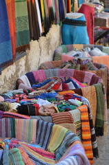 Colorful textiles and scarves displayed on street stalls in Essaouira, Morocco, showcasing a vibrant selection of fabrics in various patterns and textures against a rustic wall
