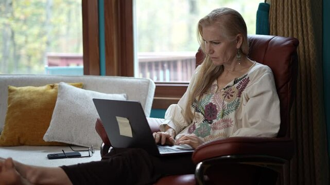 Mature woman with long blonde hair sits in a brown leather armchair, holding her forehead with her hand, looking stressed while working on a laptop. 