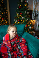 Little girl sitting wrapped in a warm red blanket against the background of a Christmas tree with red golden toys and a glowing garland at home