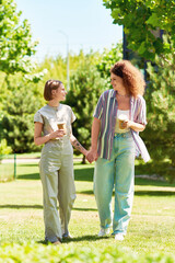 Two friends happily stroll together in a green park, sharing drinks and laughter under the sun.