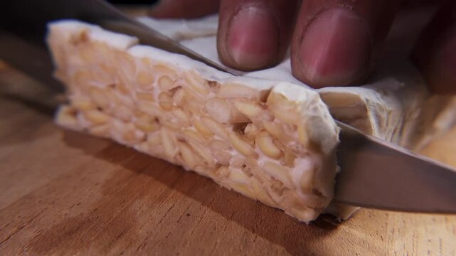 Male hand slicing tempeh with a knife on cutting board. Tempe is fermented soy product originally from Indonesia. Macro footage of vegetables