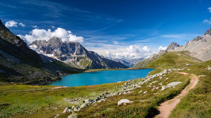 Alpine Landschaft mit kristallklarem, türkisfarbenem Bergsee, umgeben von schroffen Bergen und grünem Tal. Ein schmaler Wanderpfad führt entlang des Sees unter einem strahlend blauen Himmel.