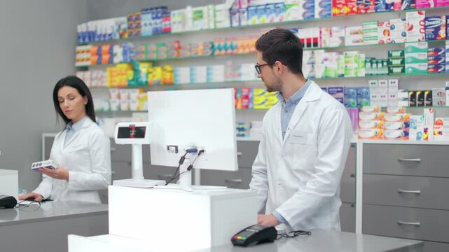 Pharmacists working with cash register. Two druggists in uniform standing near showcase with medicine. Man and woman talking in pharmacy. Male and female introducing new batch of drugs into system.