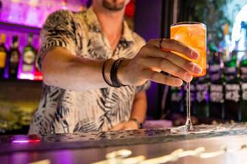 A hand holding a glass of orange beverage with a blurred bar background. The drink is garnished with a slice of orange. The scene suggests a casual, social atmosphere in a bar or restaurant.