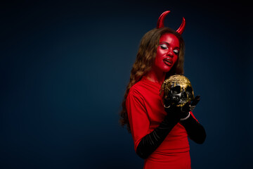 A stunning woman dressed as a devil embraces a decorative skull, exuding Halloween spirit.