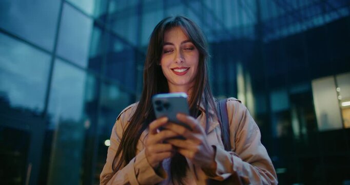 Evening in city. Cute Caucasian girl finishing entering text on her smartphone. Lowering mobile device while glancing at camera. Smiling with joy. Tall building with glass windows in background.