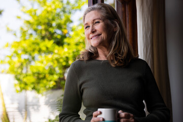 Smiling senior woman enjoying coffee by window, feeling relaxed at home