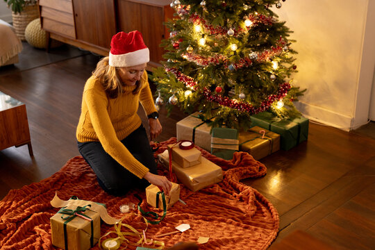 senior woman wrapping gifts by Christmas tree at home, wearing Santa hat