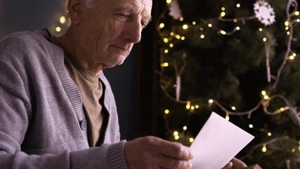 A retired man cries while looking through photos in a photo album, remembering his youth and childhood while sitting near a Christmas tree on the eve of the holiday