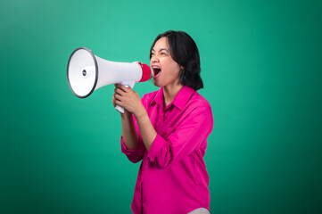 An Asian woman in a bright pink shirt against a green background, enthusiastically speaking into a megaphone with an expressive, animated face, conveying a sense of urgency or loud announcement.