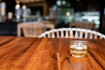 A cozy scene featuring a lit candle in a glass holder, set on a wooden table with soft, blurred background lighting, creating a warm and inviting atmosphere.