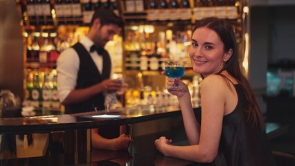 Sitting beautiful woman guest looking at camera to pose with drinking mixed vodka in blue hawaii cocktail at luxurious counter bar for Friday meeting on smart bartender background at club. Vinosity.