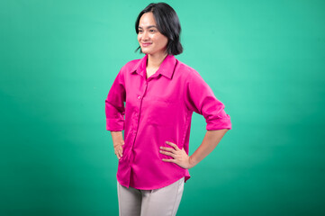 An Asian woman in a bright pink button-up shirt stands confidently with her hands on her hips against a solid green background, projecting a friendly and assertive expression