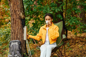 In a colorful autumn forest, a young woman relaxes with a warm drink, surrounded by vibrant foliage.