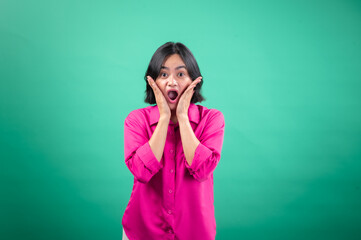 An Asian woman in a vibrant pink shirt stands against a green background, expressing surprise with her hands on her cheeks and an open-mouthed smile, capturing a lively and joyful reaction