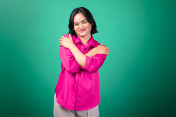 An Asian woman in a pink shirt stands against a green background, crossing her arms over her chest in a self-hug gesture, expressing warmth, self-love, and comfort with a gentle smile