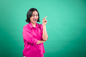 An Asian woman in a vibrant pink shirt stands against a green background, looking cheerful and pointing upwards with one hand, as if indicating a bright idea or something exciting
